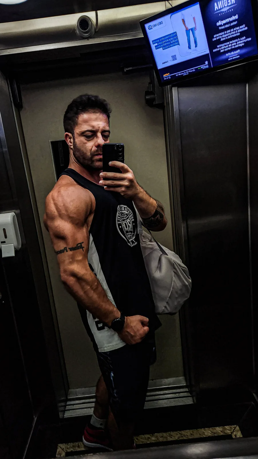Montinho Personal de regata preta e mochila no ombro, em selfie no espelho do elevador a caminho de um atendimento — personal trainer destacado pelo portal para quem treina em Cajamar