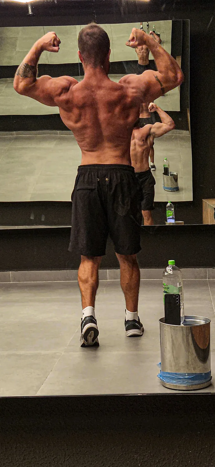 Montinho Personal de costas em pose de duplo bíceps diante do espelho da academia, mostrando as costas e os ombros definidos — personal trainer destacado pelo portal para quem treina em São Roque