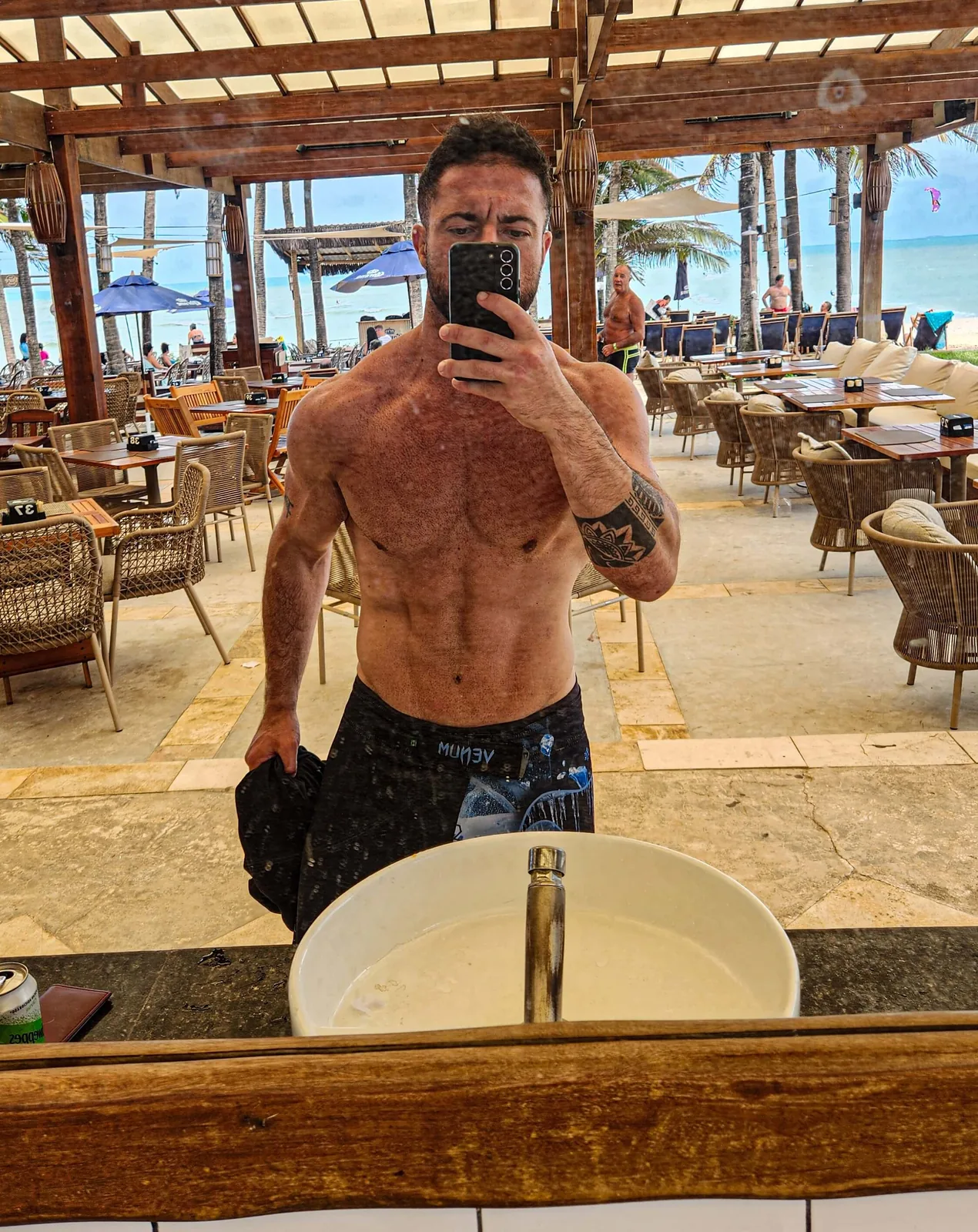 Montinho Personal sem camisa em selfie no espelho de um quiosque à beira-mar, com a praia ao fundo — personal trainer destacado pelo portal para quem treina em Itapevi